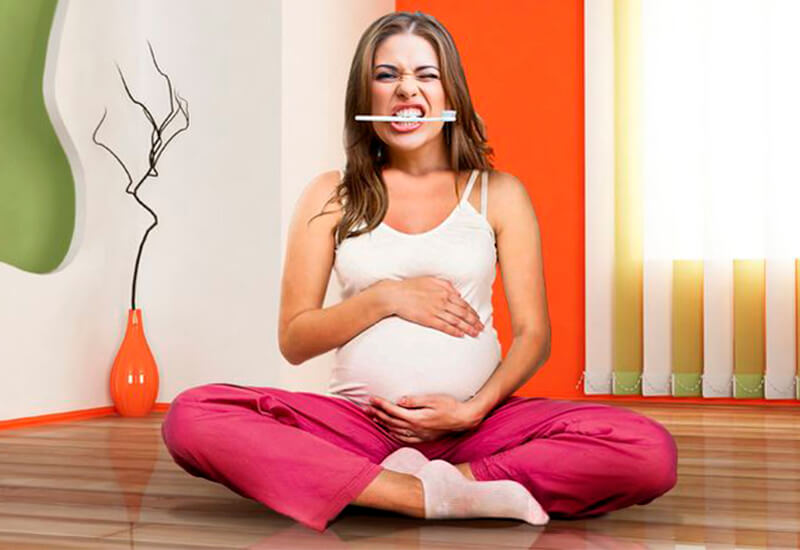 Беременная женщина сидит на полу, в зубах держит щетку